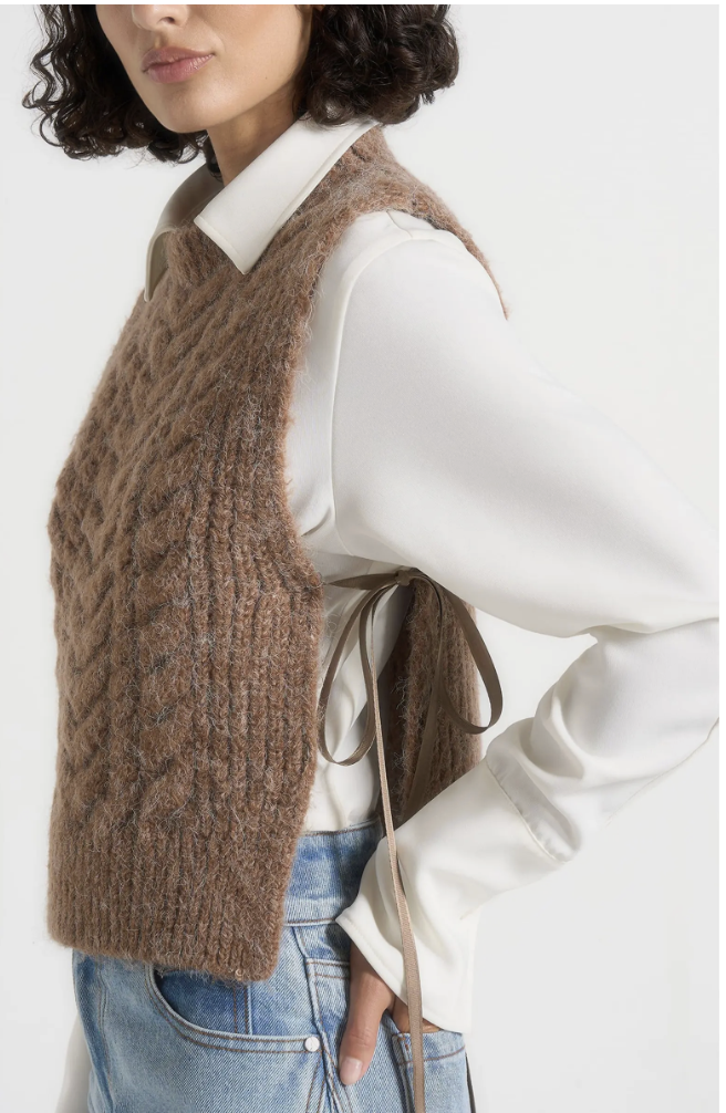 A side view of a woman wearing a brown wool cable-knit sweater vest with side tie closures, layered over a white long-sleeve top and paired with light wash jeans.
