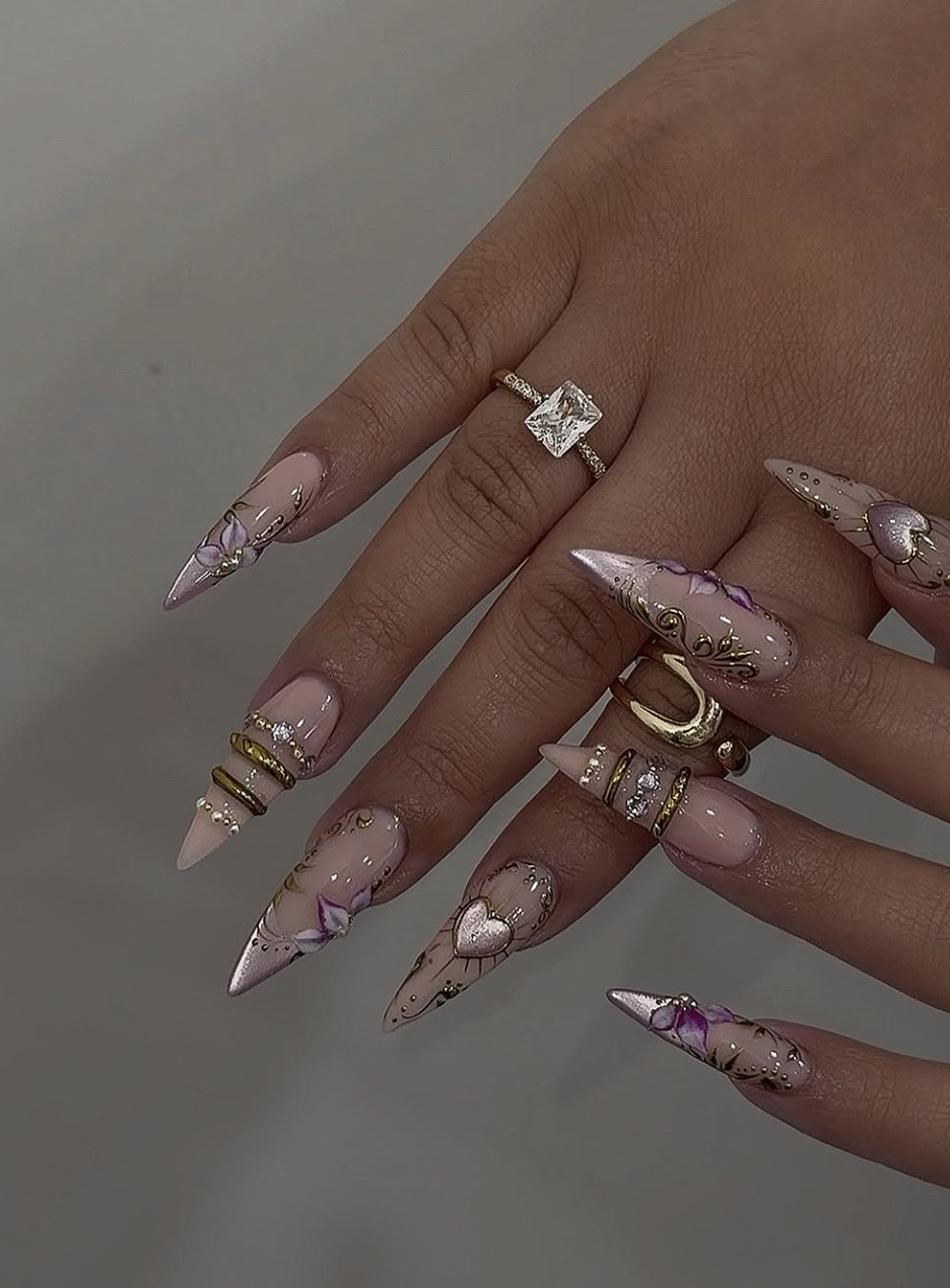 Nude almond nails featuring 3D gold heart and star charms, textured gold bangles, and delicate purple floral accents.