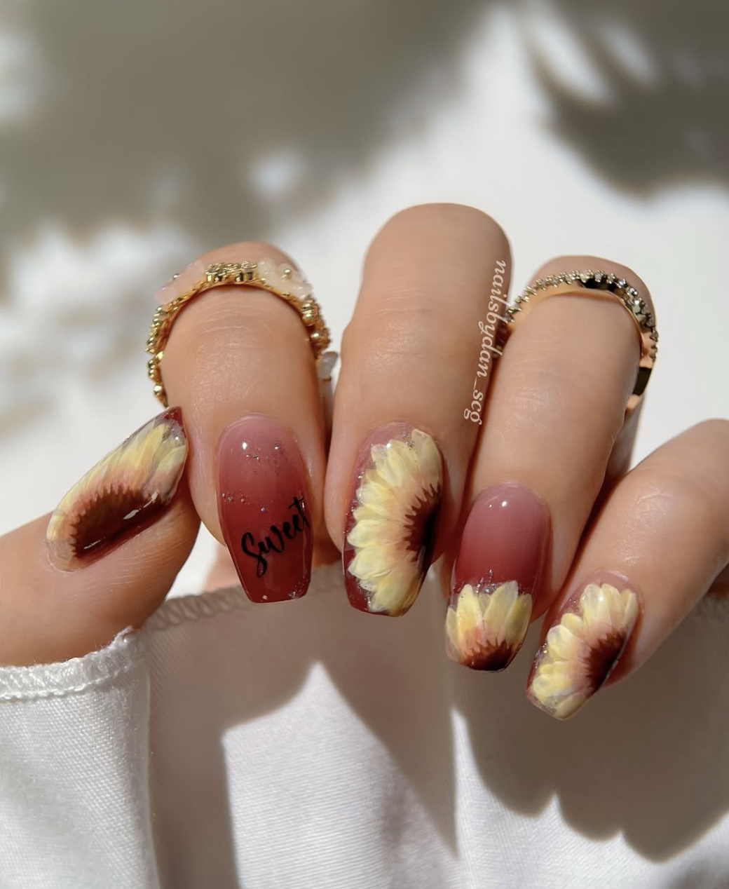 Long coffin-shaped nails with a deep burgundy-red base, featuring large hand-painted sunflowers and the word "Sweet" written in elegant black script on the middle finger.
