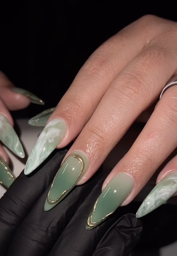 A high-fashion matcha green nail art design featuring a mix of solid green, white marble patterns, and elegant gold metallic leaf accents on long almond-shaped nails