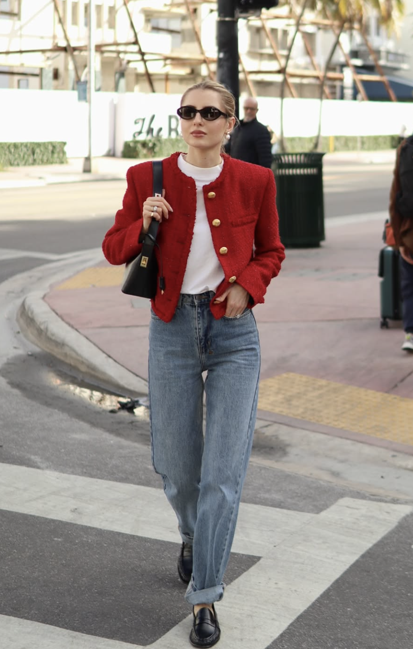 A woman wearing a structured red button-down lady jacket with gold buttons, a white t-shirt, and medium-wash slim-straight jeans walking on a city street.