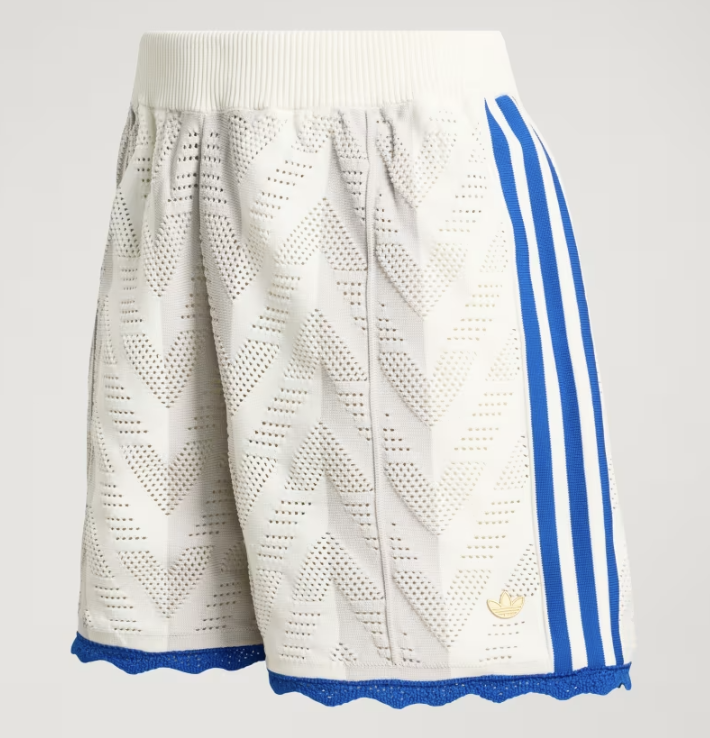 A close-up of white Adidas Originals knit track shorts featuring a textured chevron pattern, royal blue side stripes, and a scalloped blue lace hem.