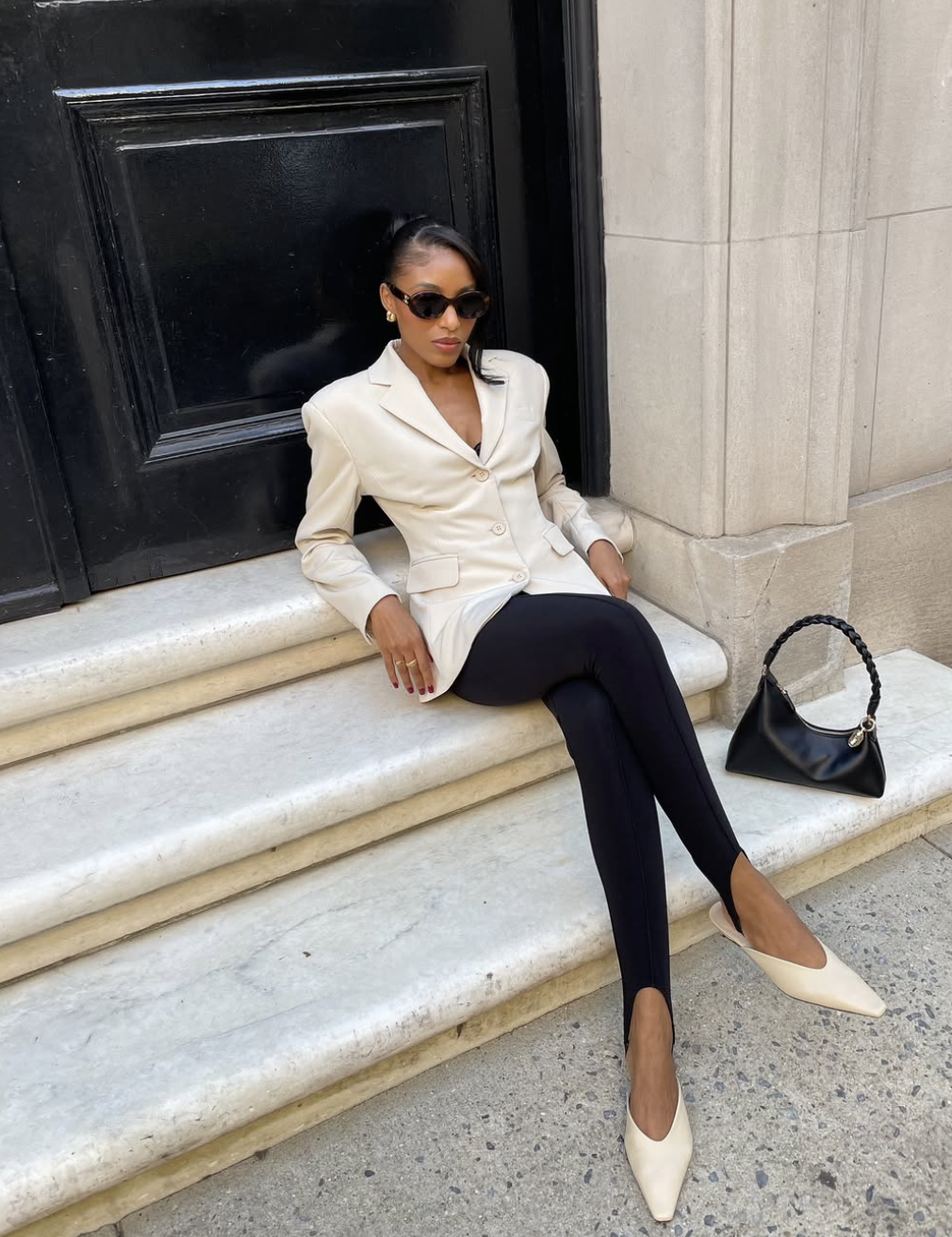 A woman sitting on stone steps wearing a structured cream blazer over black stirrup leggings and white pointed-toe flats.