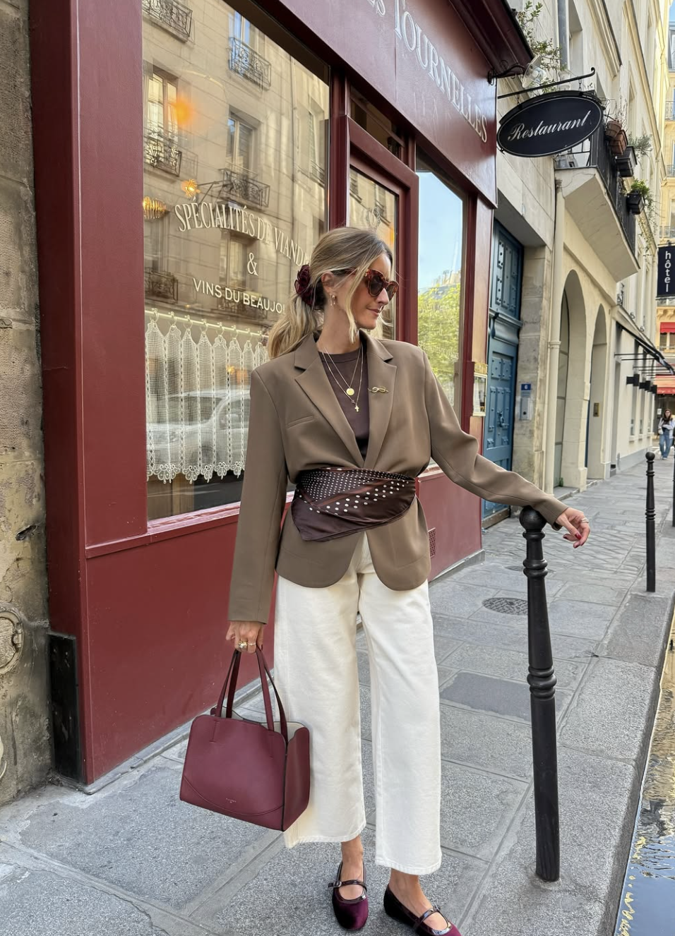 A woman wearing an oversized cappuccino-colored blazer cinched at the waist with a patterned silk scarf belt, styled with dark trousers and a black leather bag.