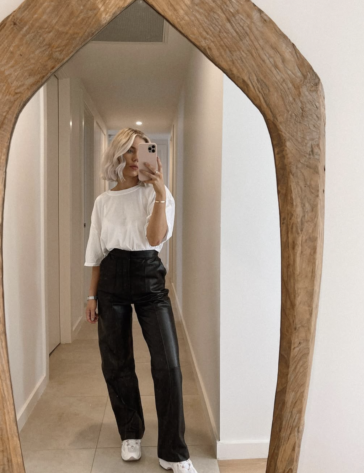 Woman wearing black straight-leg leather pants with a white t-shirt and white sneakers in a mirror selfie