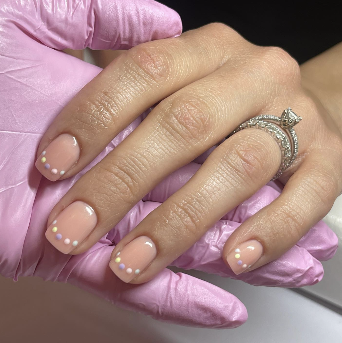 A close-up of a hand with short square-shaped natural nails featuring a sheer nude base and three tiny, vertical pastel dots in yellow, white, and mint green on the side of each nail.