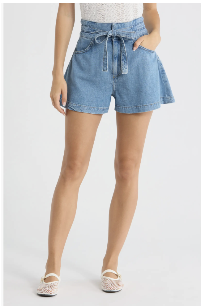 A close-up of a woman styling high-waisted denim paperbag shorts with a cinched tie belt, paired with a white top and clean white slides.