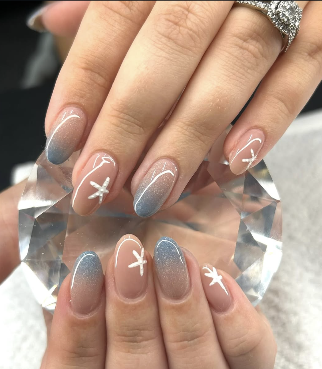Oval-shaped nails featuring a soft nude base that fades into a glittery blue ocean ombre tip, accented with hand-painted white starfish, highlighting cute June nail ideas.