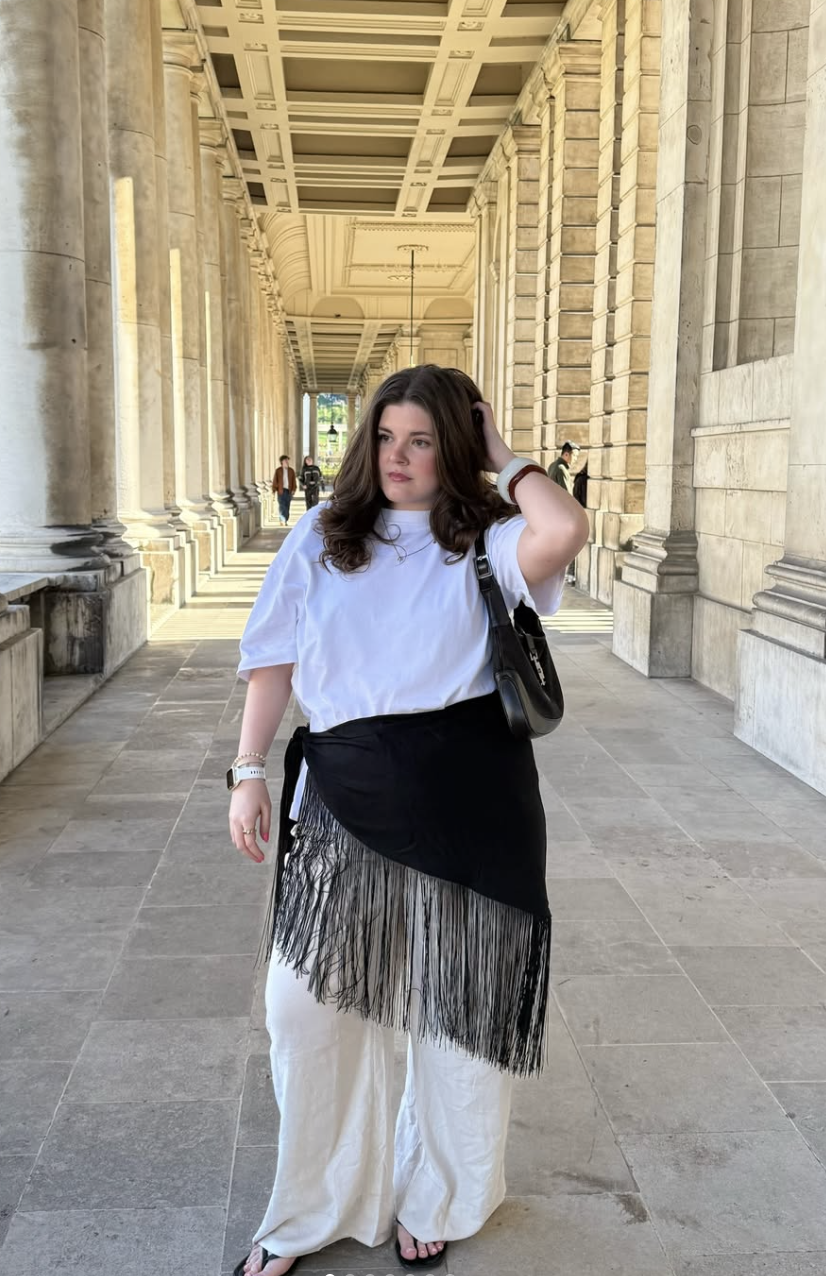 A street style shot of a woman standing between classic architectural columns, wearing a large black scarf with long fringe detailing styled as a wrap skirt over high-waisted white trousers and a casual white t-shirt.