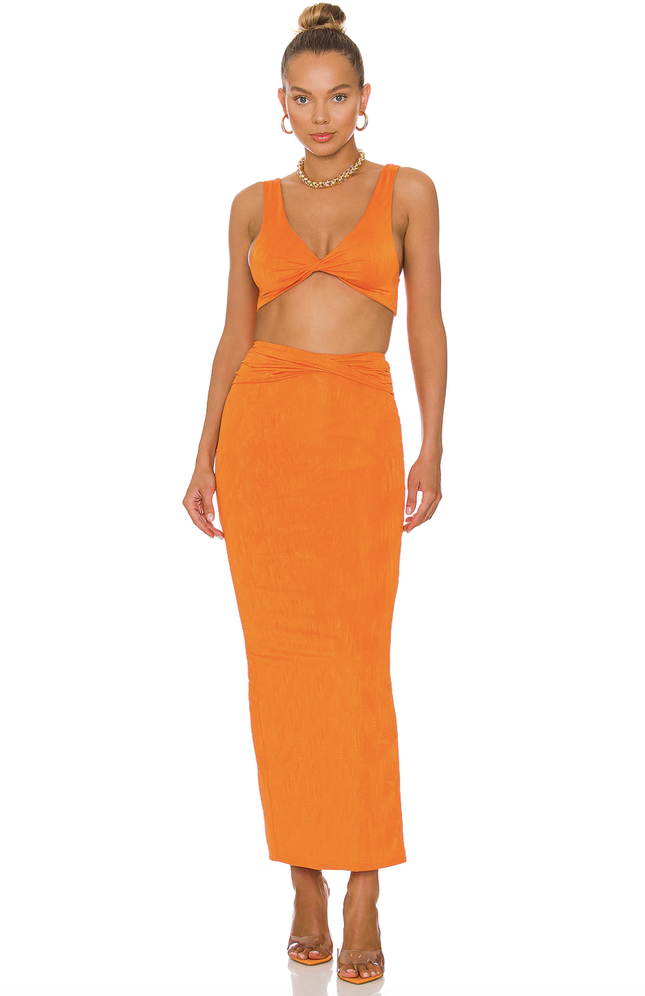 A woman with her hair styled in a high bun wearing a vibrant orange two-piece set, featuring a twisted sleeveless crop top and a matching high-waisted maxi skirt, accessorized with a gold chain necklace and gold hoop earrings.