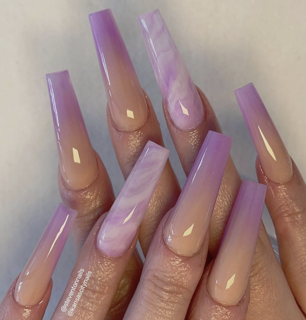 Long stiletto-shaped nails featuring a seamless ombre fade from white to lavender with delicate purple marble accents.