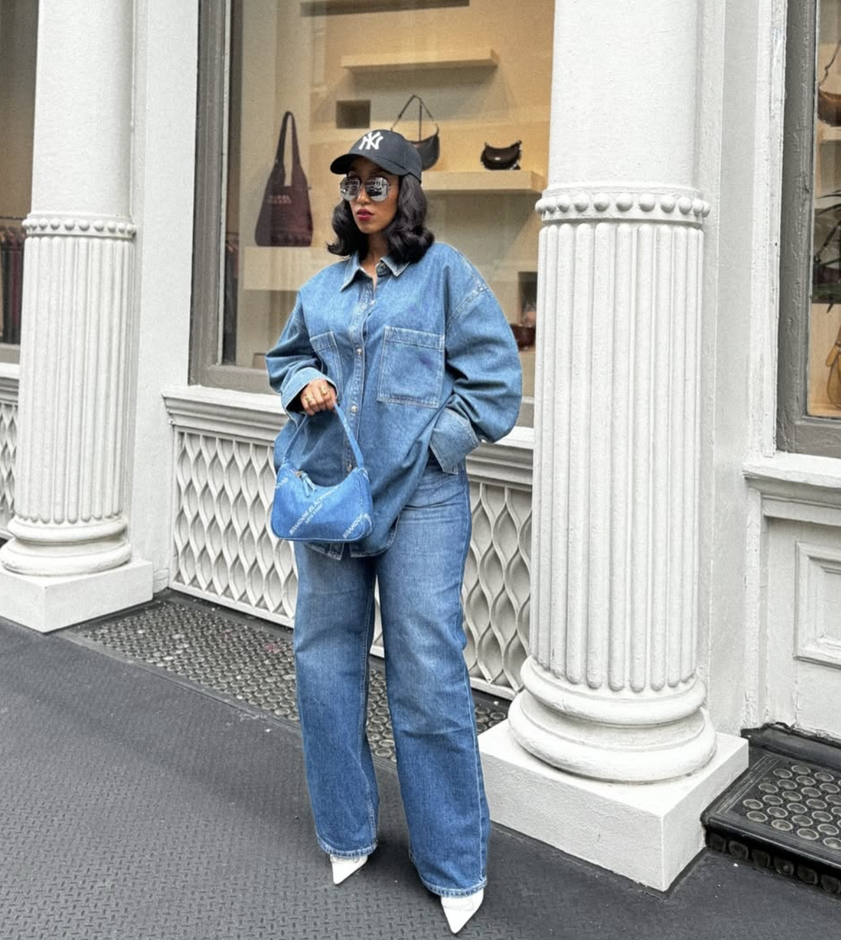 A woman wearing a matching light blue denim set featuring an oversized denim button-down shirt tucked into wide-leg jeans, styled with a blue quilted handbag, white pointed-toe boots, and a black baseball cap.