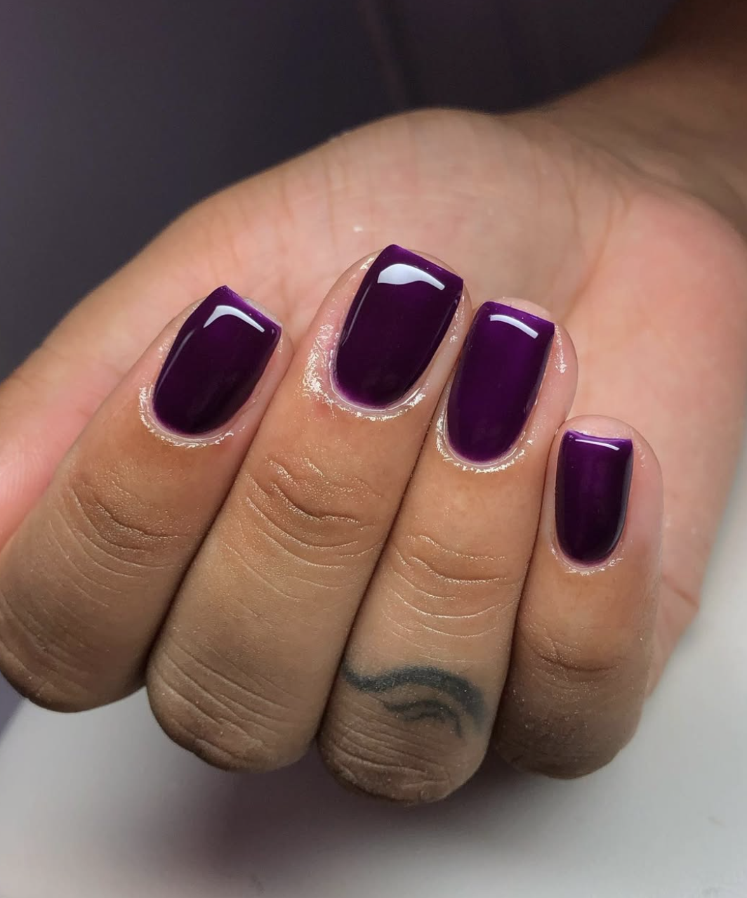 Close-up of short nails featuring a glossy dark plum manicure with a small finger tattoo.