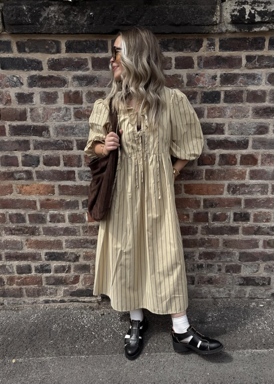A woman wearing a beige and black striped puff-sleeve midi dress paired with thick white crew socks and chunky black T-strap fisherman sandals, holding a brown suede tote bag against a brick wall.