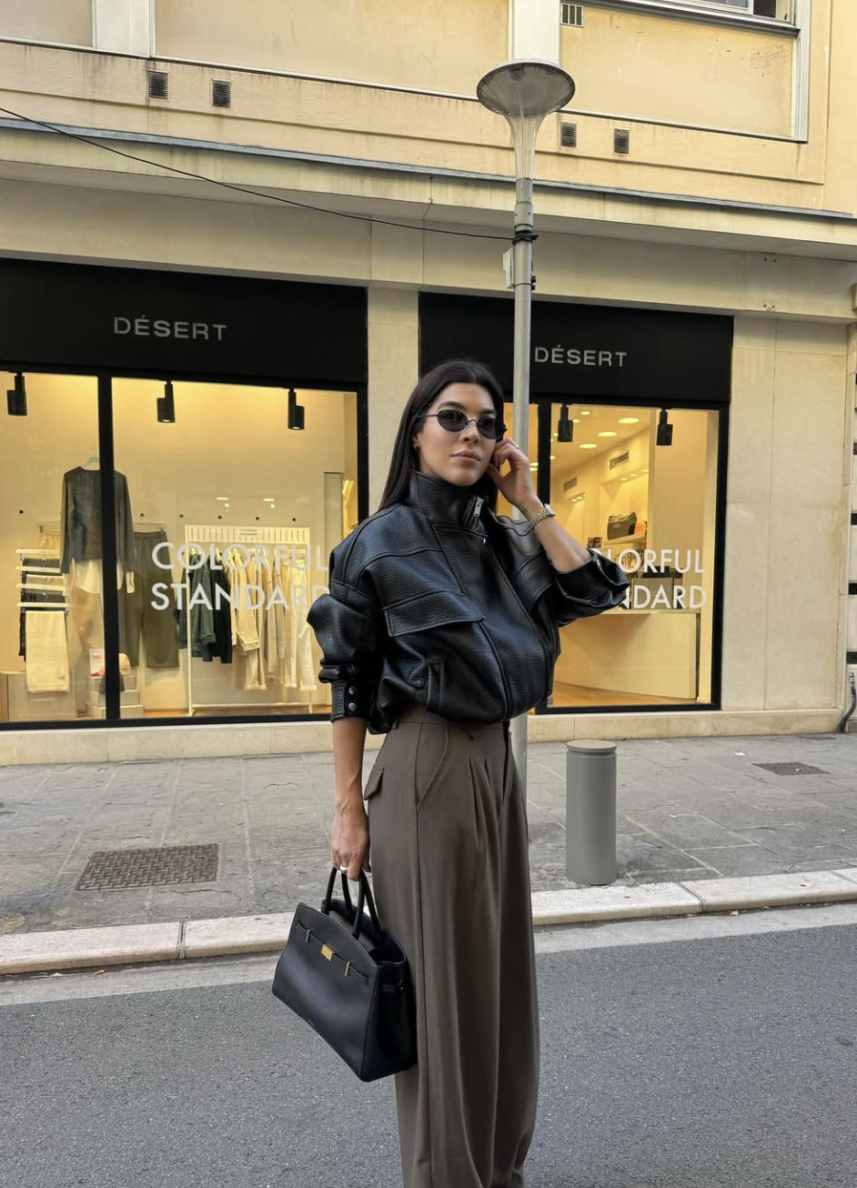 A woman wearing a modern utility-inspired neutral outfit featuring a cropped black leather bomber jacket tucked into high-waisted taupe wide-leg trousers, paired with a structured black handbag.