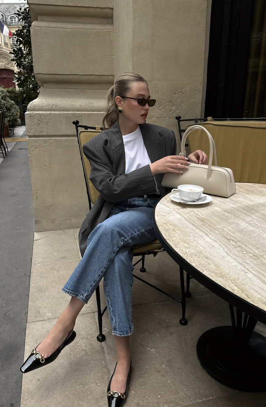 Close-up of a person wearing black pointed-toe ballet flats with gold chain detail paired with slim-straight light wash jeans while sitting at an outdoor cafe.