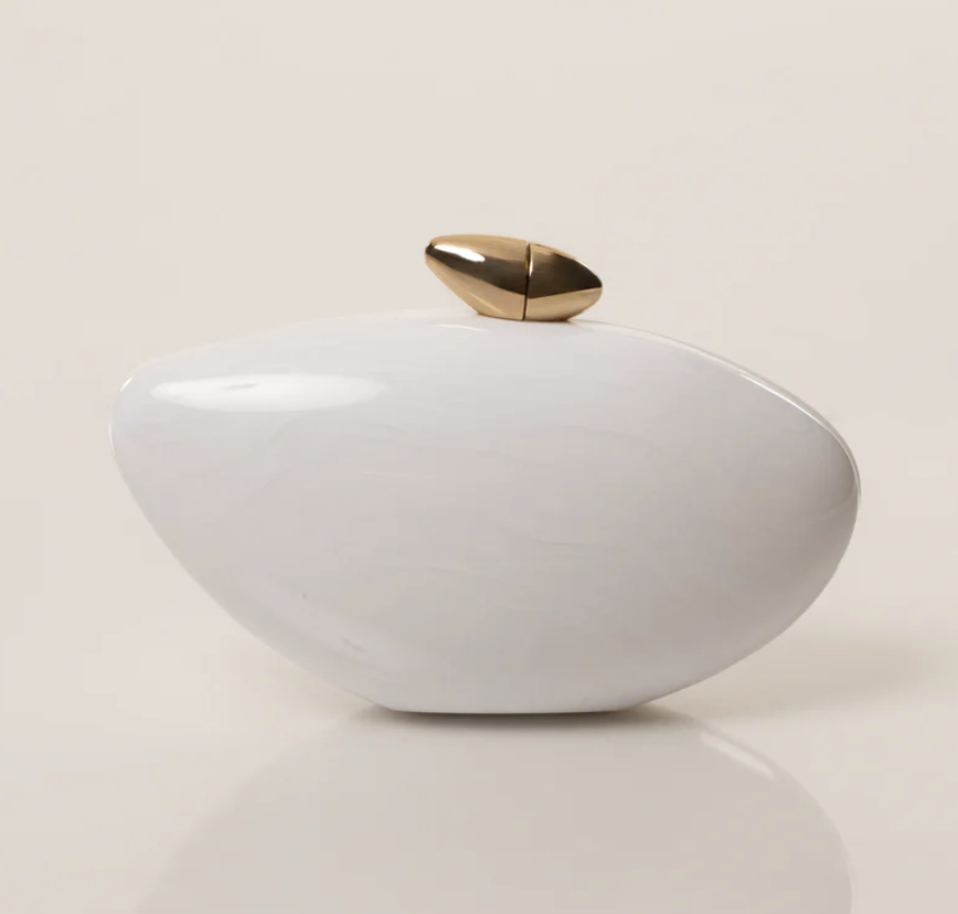 An organic-shaped white hard-shell clutch bag with a large gold and pearl top clasp, styled as a luxury accessory for a Mediterranean resort evening look.