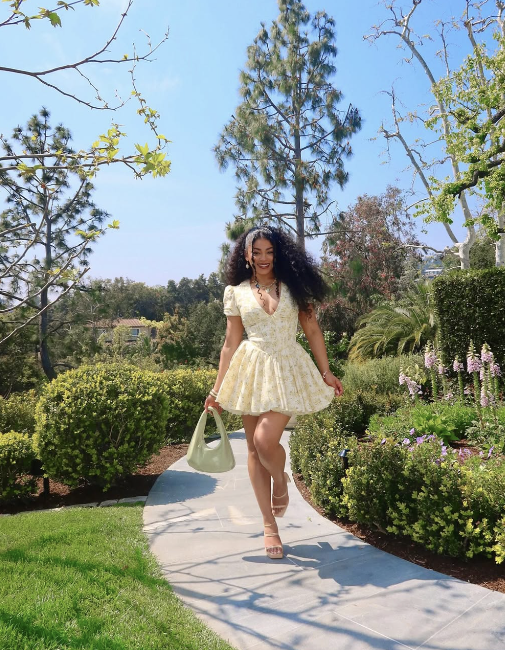 A woman walking outdoors in a light-colored floral mini dress with a matching pastel green handbag, perfect for a 2026 Easter brunch.