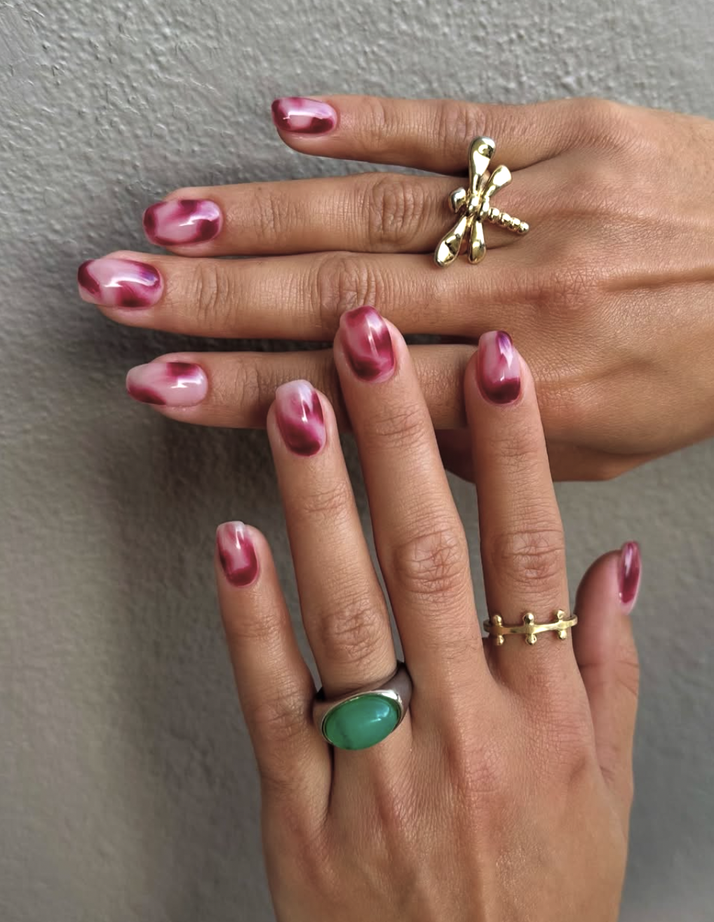 A close-up of short natural nails featuring vibrant hot pink manicure accents, styled with gold and green gemstone rings.