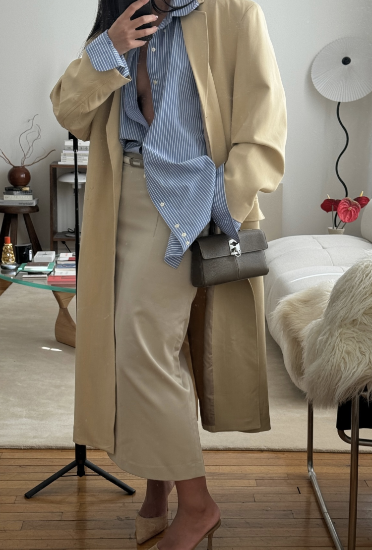 A chic spring outfit featuring a classic tan trench coat layered over a blue striped button-down shirt and beige wide-leg trousers, styled with a small dark crossbody bag.