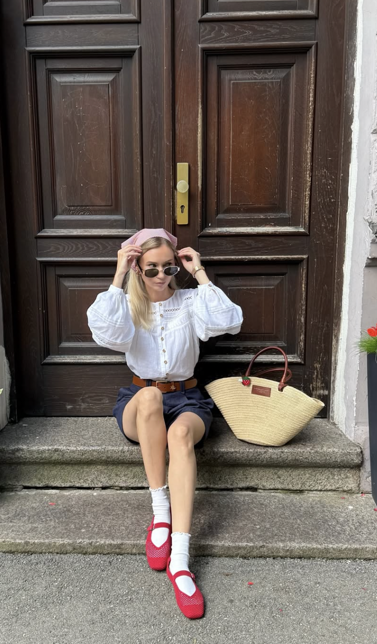 A woman sitting on stone steps wearing a pink headscarf, a white puff-sleeve blouse, navy shorts, white ruffle ankle socks, and red ballet flats, next to a large straw tote bag.