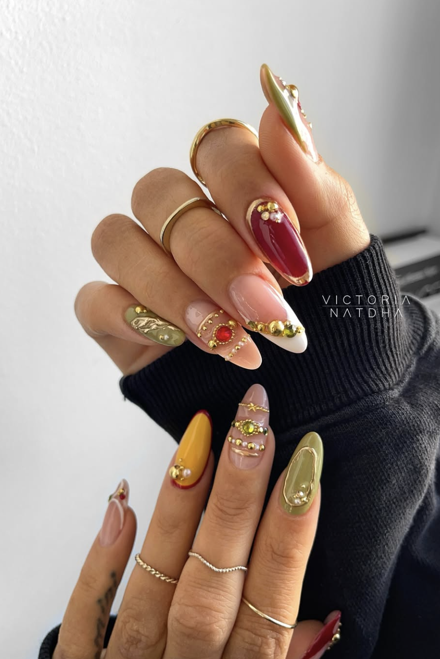Long almond nails with a mix of deep ruby red and amber yellow polish, featuring ornate 3D gold bands and large red gemstone charms.