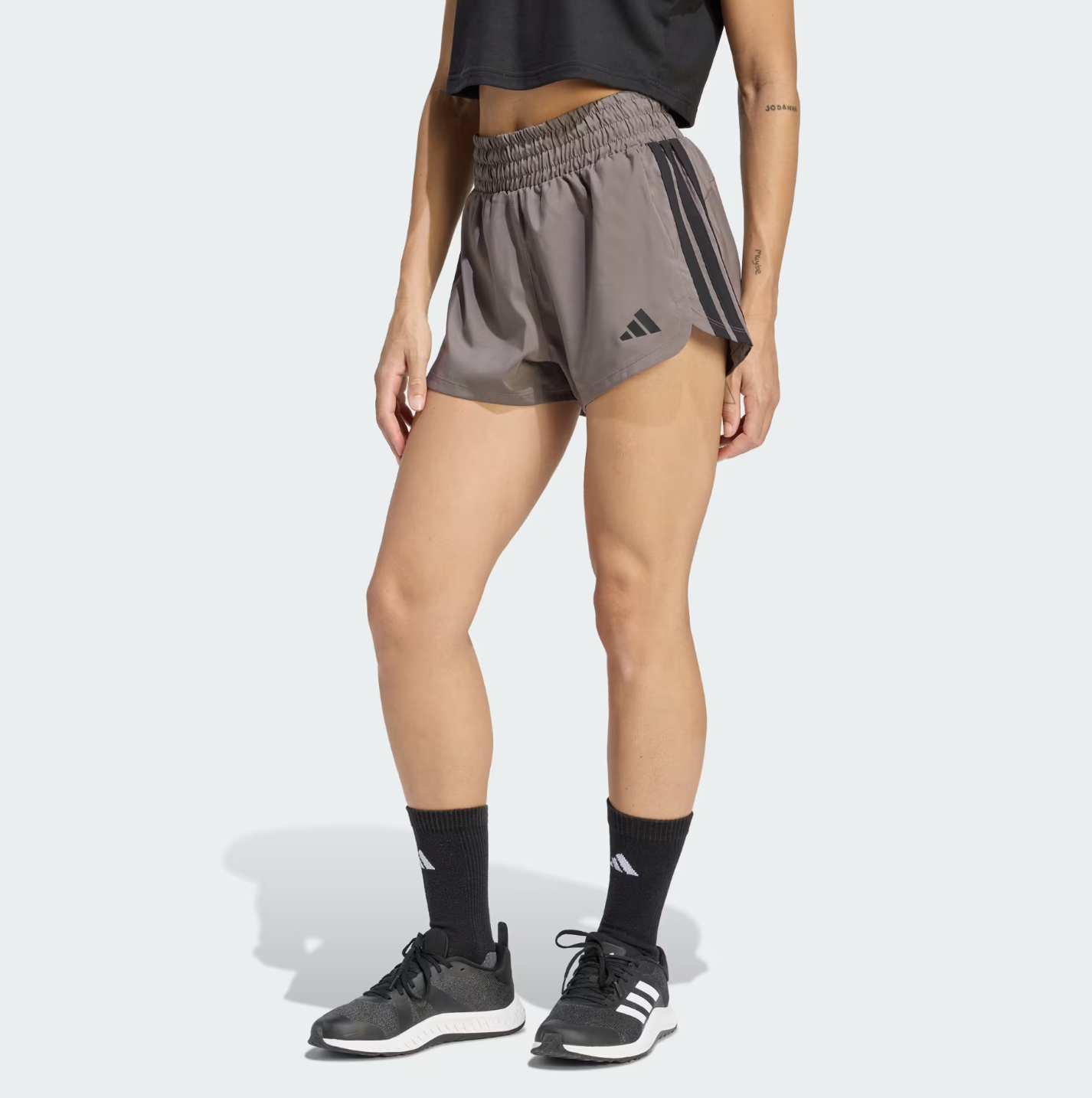 A close-up of a person wearing grey high-rise Adidas Pacer workout track shorts with signature dark grey stripes on the side, paired with black socks and black athletic sneakers.