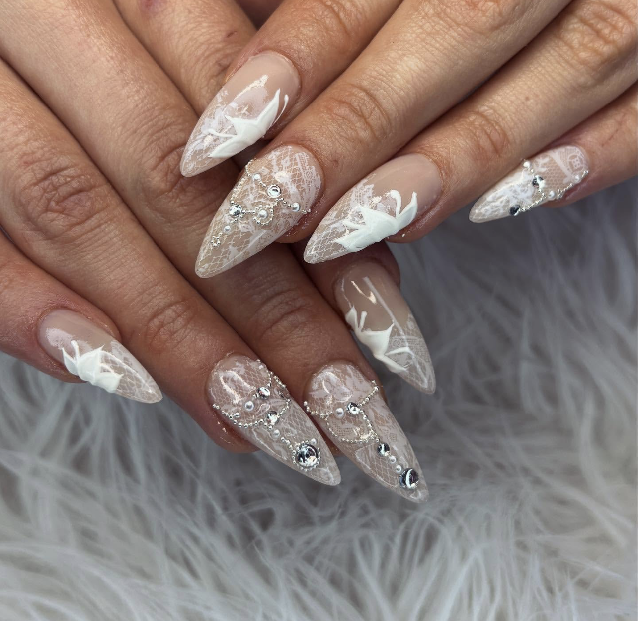 Long stiletto-shaped acrylic nails with a nude base featuring white 3D floral lace patterns, small pearls, and rhinestone accents.