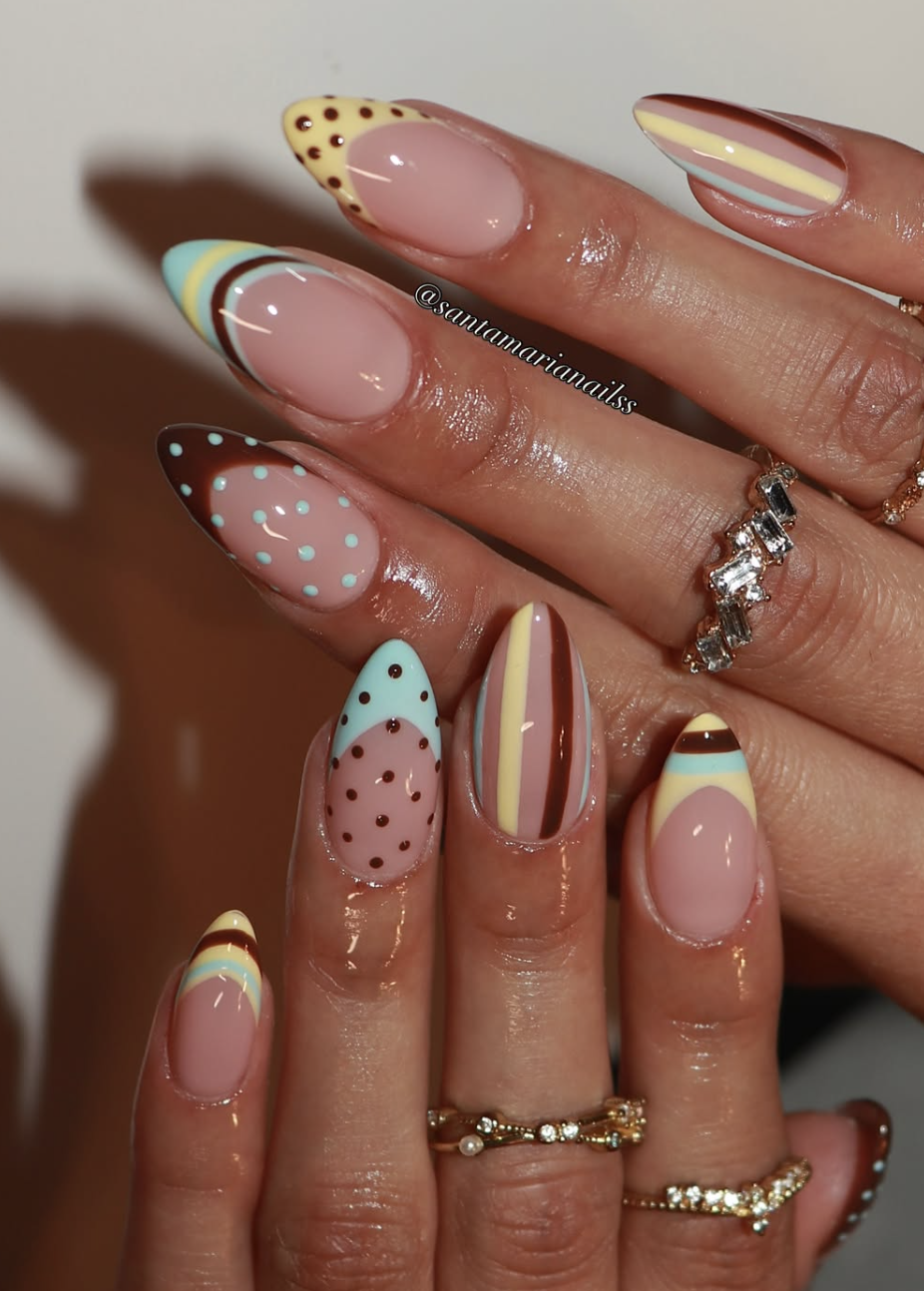 A close-up of almond-shaped nails featuring a mix of colorful French tips and full accent nails decorated with vertical stripes, polka dots, and scalloped details in pastel yellow, brown, mint green, and pink.