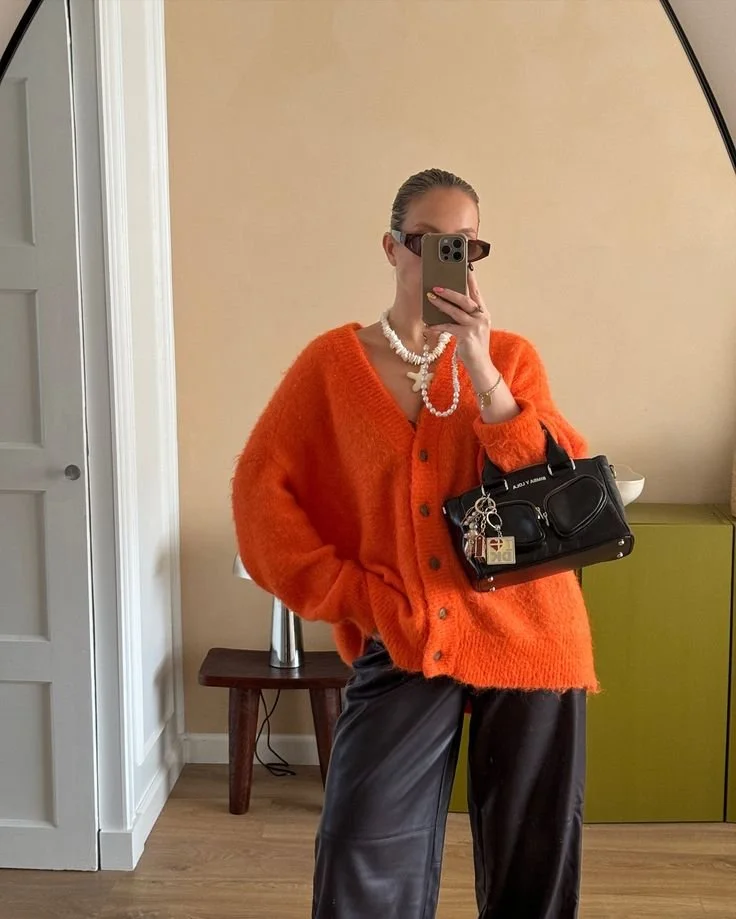 A fashion editor-approved styling for Spring 2026, featuring a vibrant Persimmon orange knit cardigan paired with black leather trousers and a quilted handbag with gold hardware.