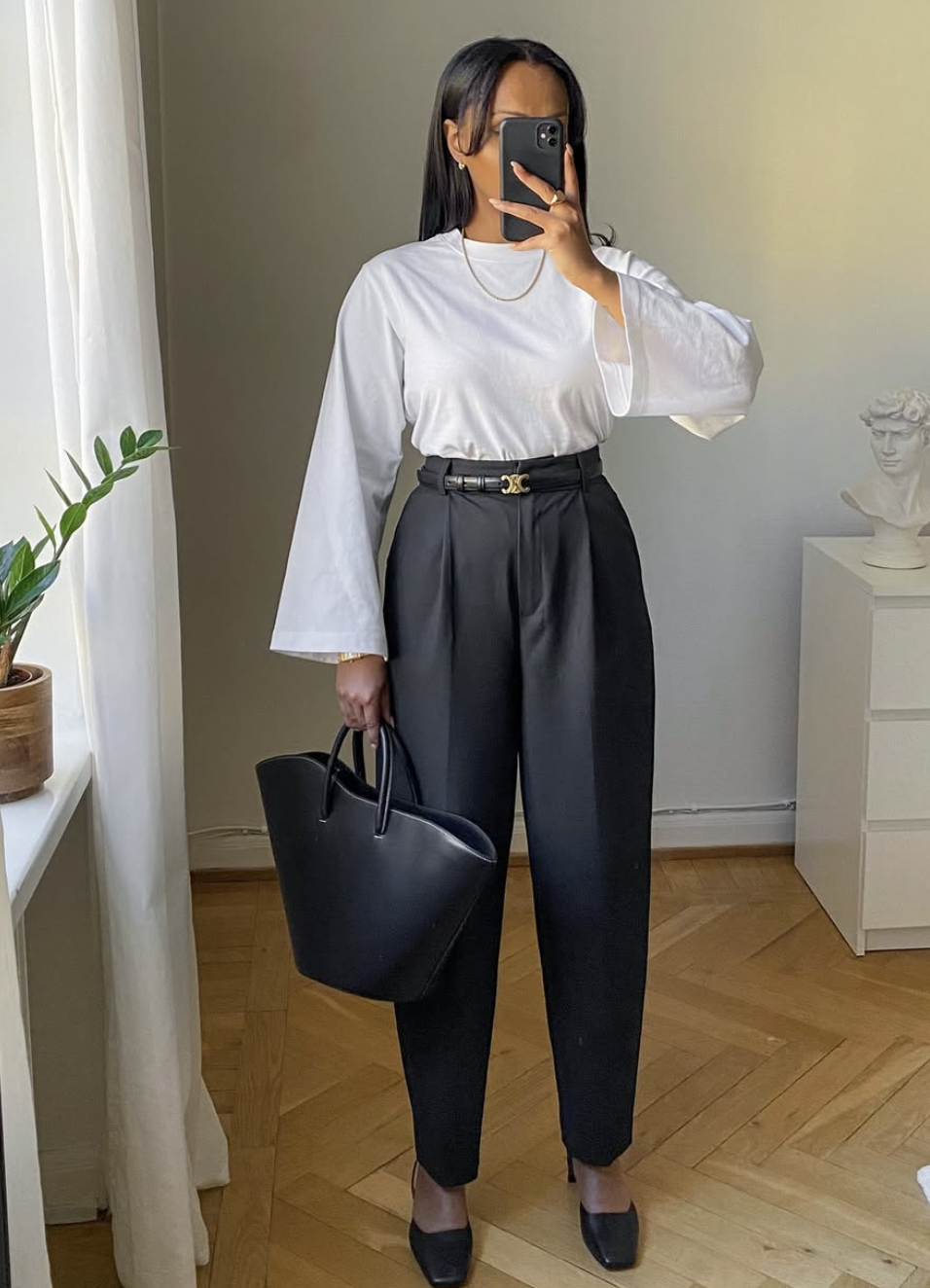 A woman wearing a white long-sleeve top tucked into high-waisted black pleated trousers with a thin belt, styled with black Mary Jane flats for women and a black leather tote bag.