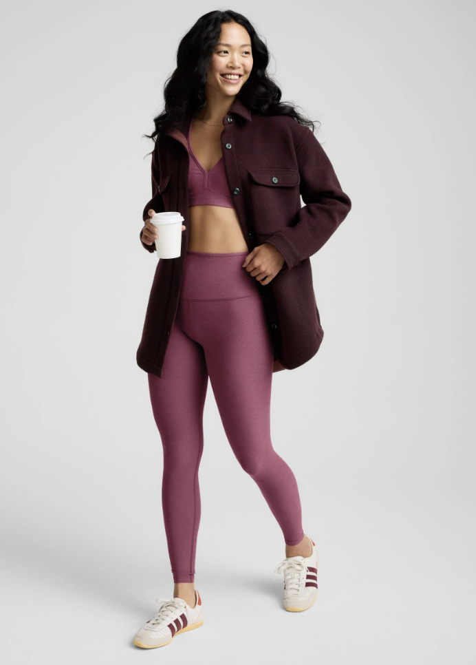 A model wearing high-waisted magenta leggings with a matching sports bra, layered under a long dark maroon shacket, holding a coffee cup to demonstrate a functional winter 2026 everyday outfit.