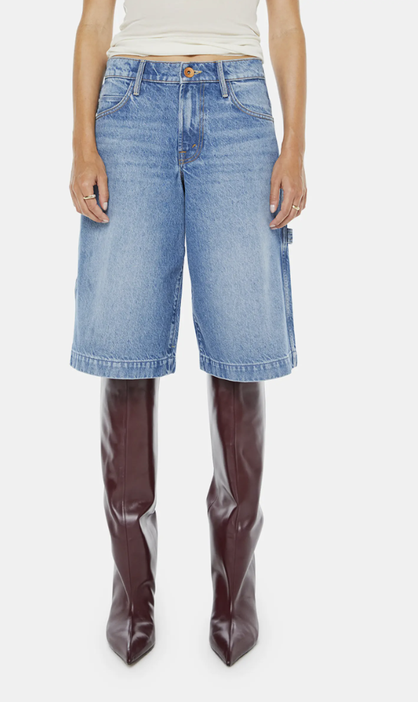 A close-up product view of a woman wearing loose-fit, light-wash denim Bermuda shorts featuring a carpenter-style hammer loop, styled with dramatic knee-high dark cherry brown leather boots.