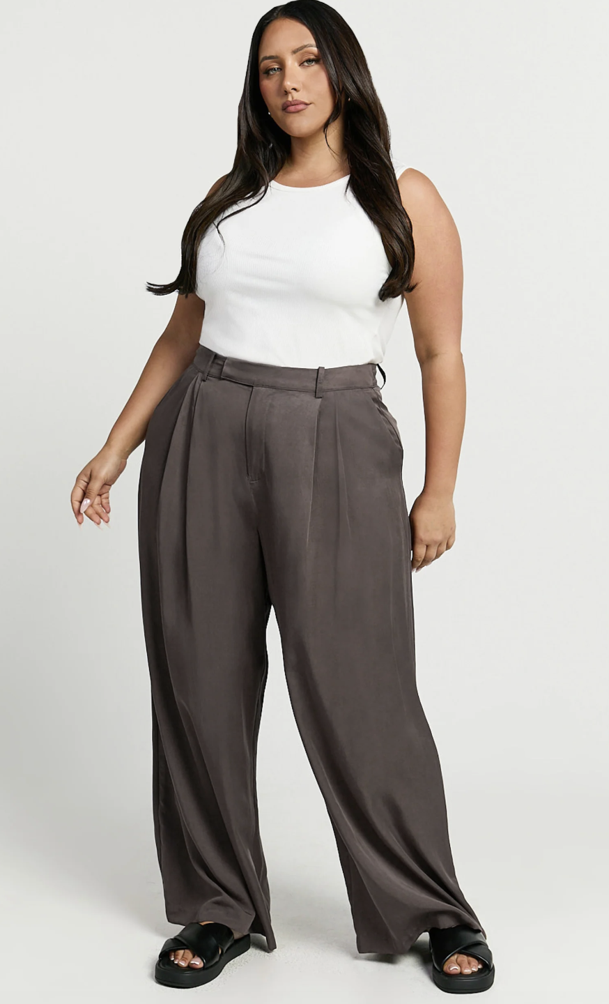 A product view of a woman wearing high-waisted, pleated wide-leg trousers in a dark charcoal grey, styled with a tucked-in white halter neck top.