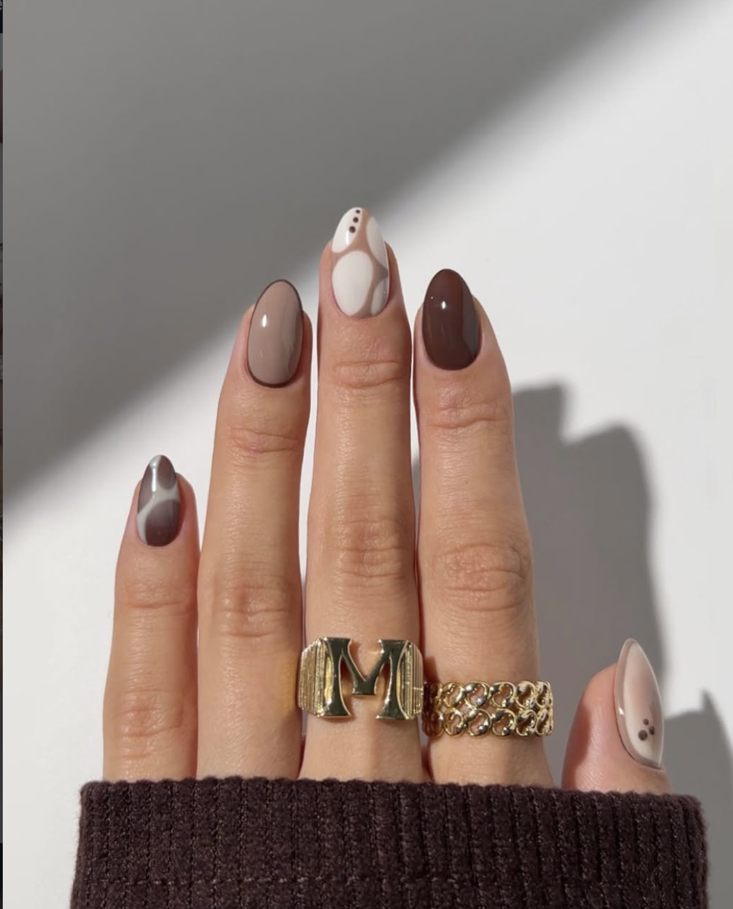 A close-up of a hand featuring a mismatched tonal brown manicure with swirl nail art, solid taupe and dark chocolate polish, and a coffee half-moon design, styled with gold rings.