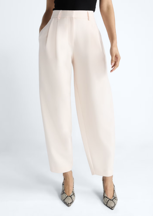 A woman wearing cream-colored high-rise pleated trousers and snakeskin pointed-toe heels from the Devil Wears Prada x Walmart collection, styled as a polished editorial wardrobe staple.