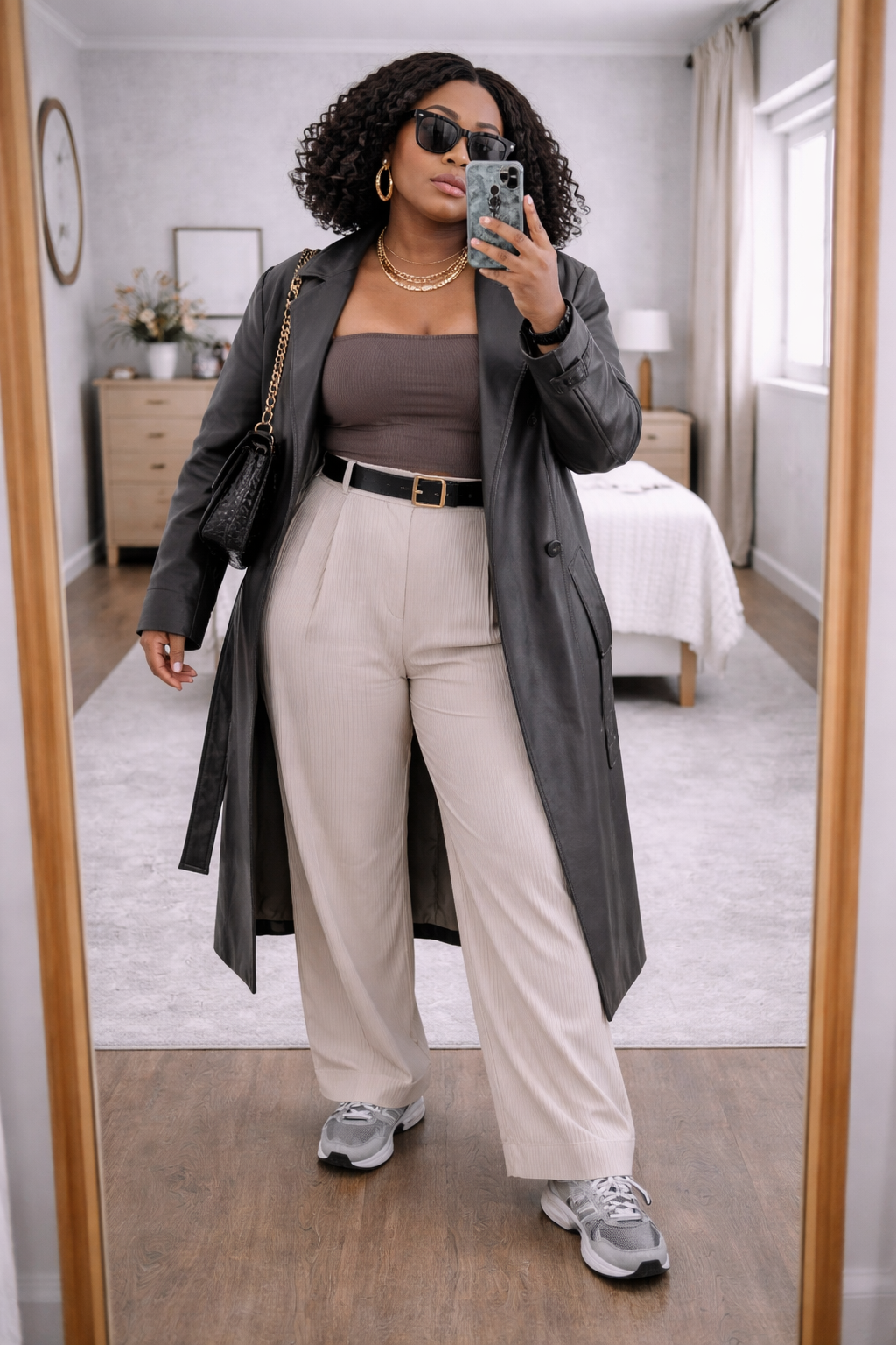 A woman taking a mirror selfie wearing a long grey duster coat over an olive green tube top and wide-leg tan trousers, accessorized with a black belt and sunglasses.