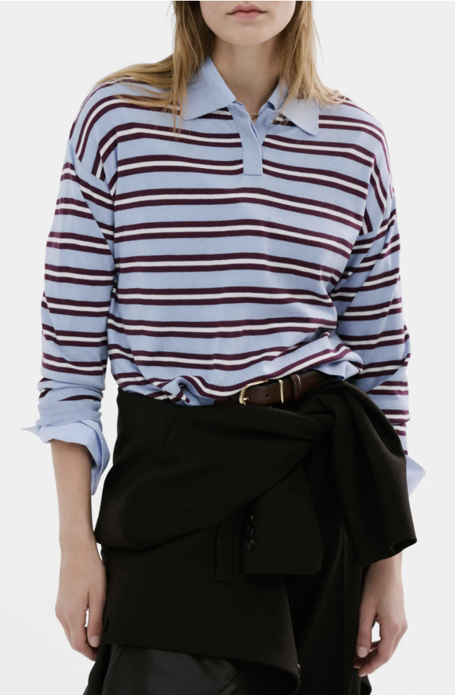 A woman wearing a light blue long-sleeve polo sweater with dark horizontal stripes, tucked into a black skirt with a brown leather belt.