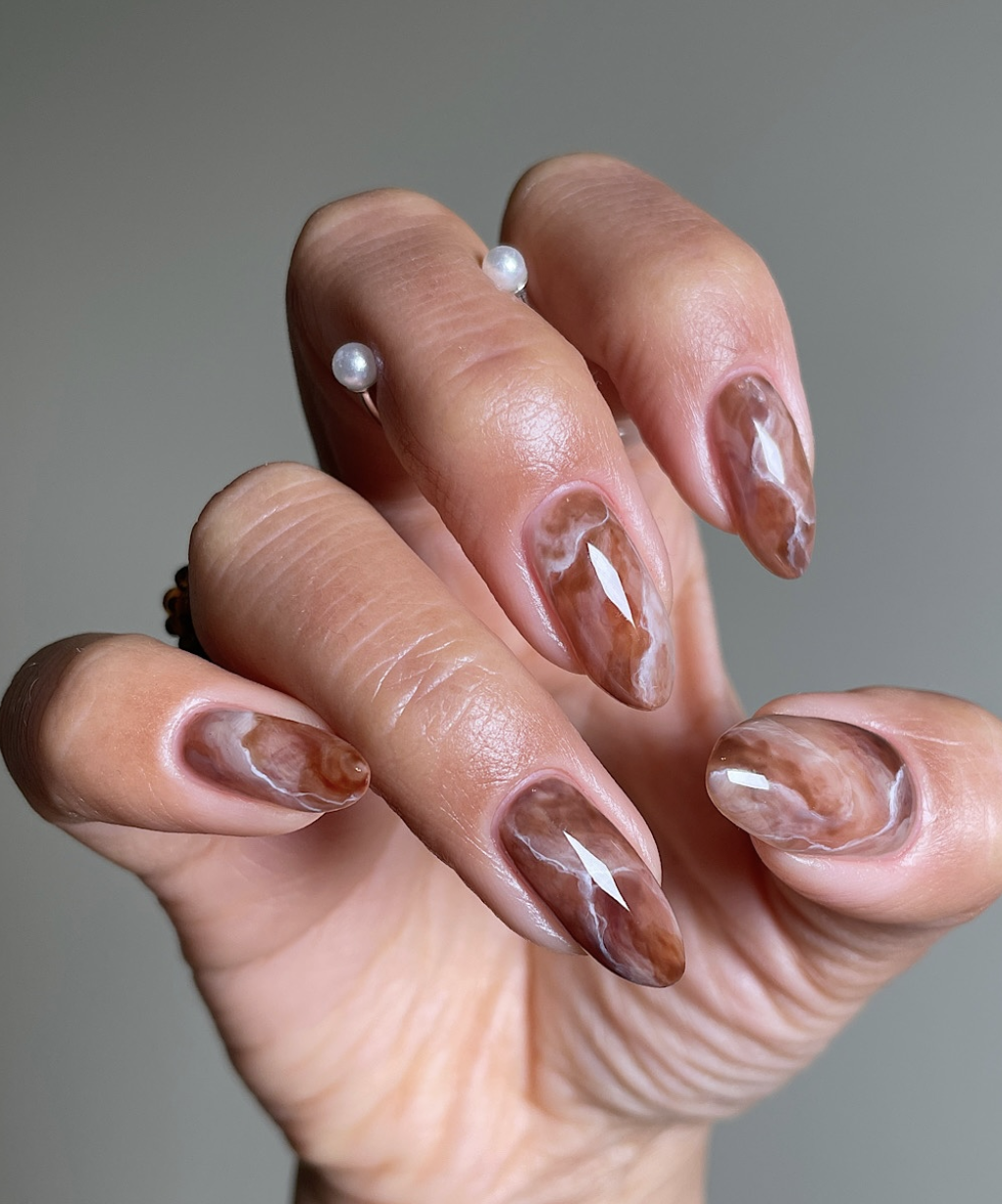 A close-up of glossy almond nails featuring abstract brown, caramel, and cream latte swirls over a sheer mocha base.