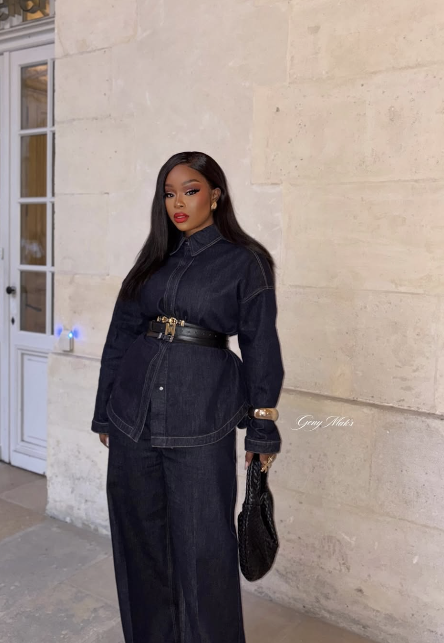 A woman wearing a dark wash denim button-down shirt belted at the waist with a black and gold belt, paired with matching dark denim jeans for a monochromatic denim-on-denim look.
