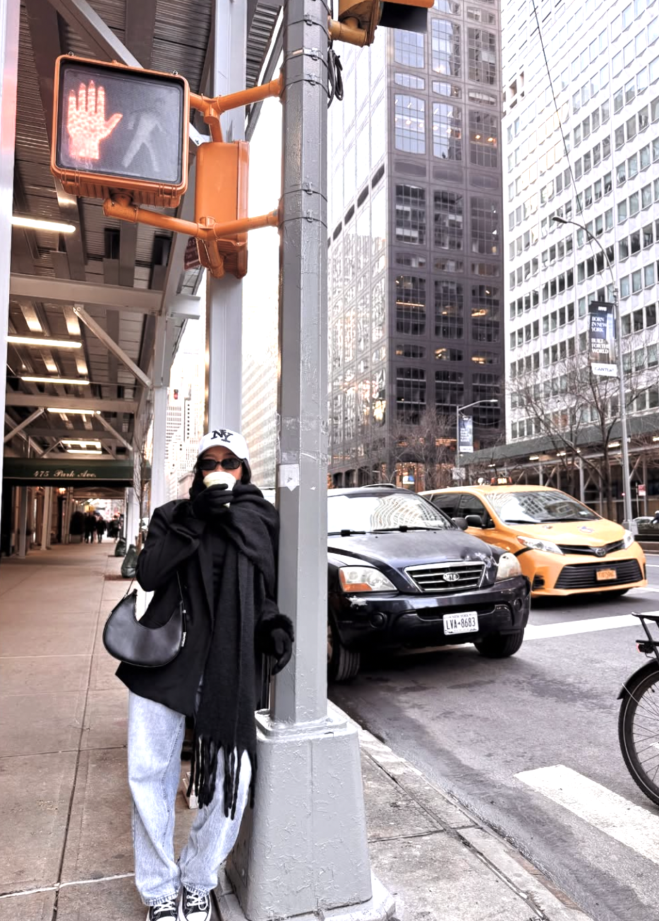 A woman wearing light-wash baggy jeans with a black oversized coat, a heavy black scarf, and a white baseball cap to demonstrate intentional layering for cold weather.