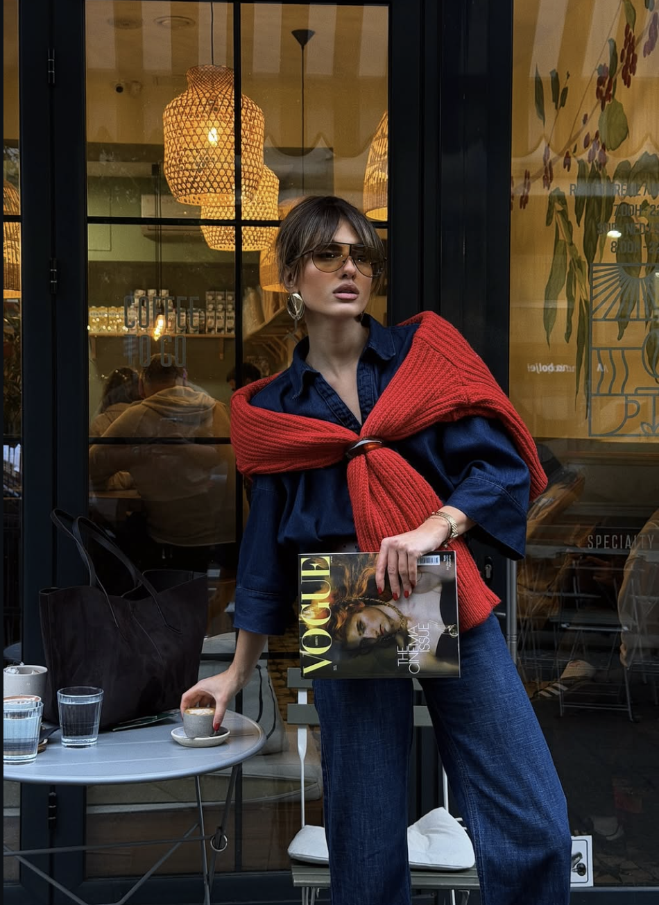 A woman wearing a denim shirt and matching jeans with a bright red knit sweater draped over her shoulders.