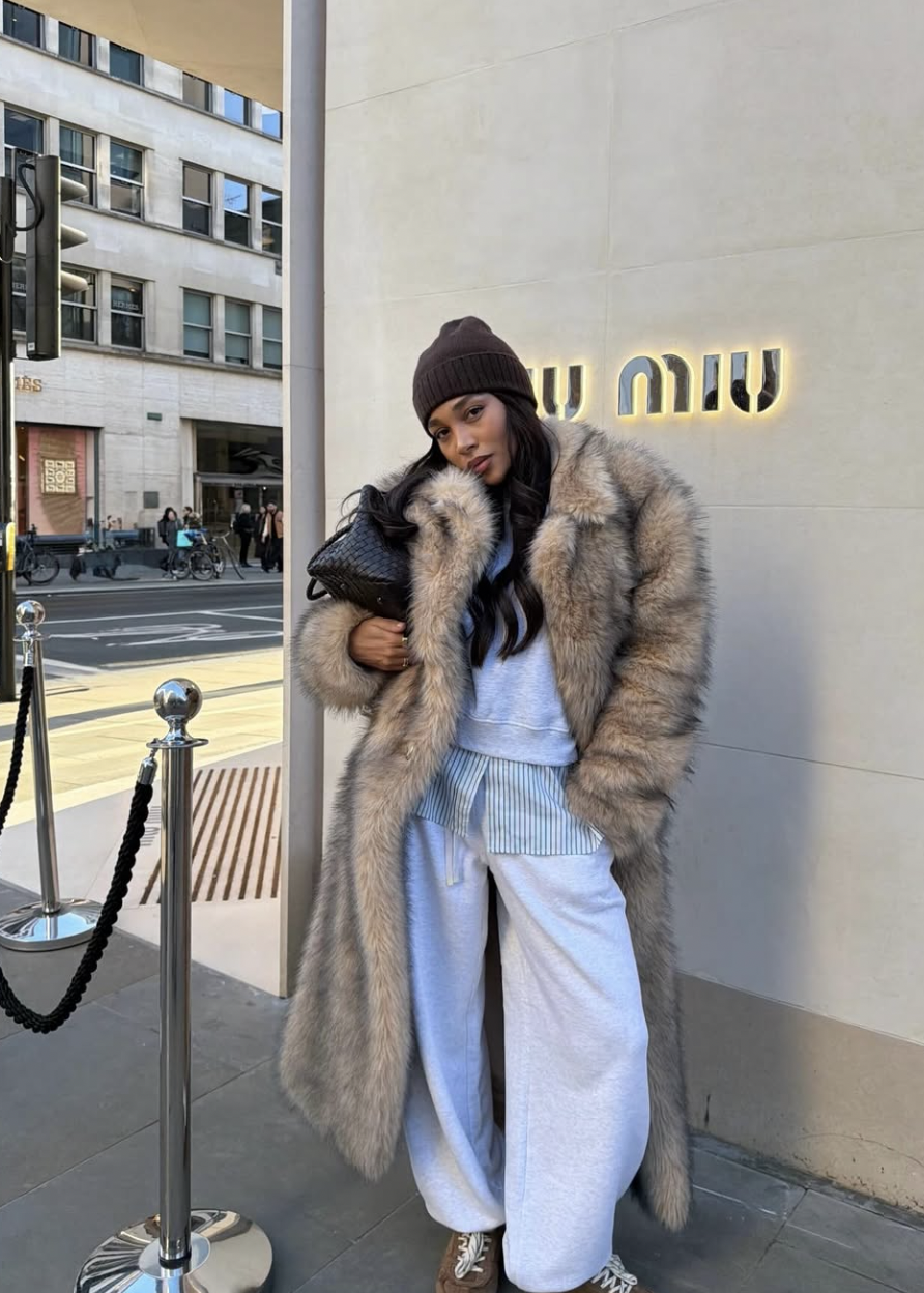 A woman wearing a cozy neutral outfit featuring a floor-length tan faux fur coat layered over a grey hooded sweatsuit with a structured black clutch.
