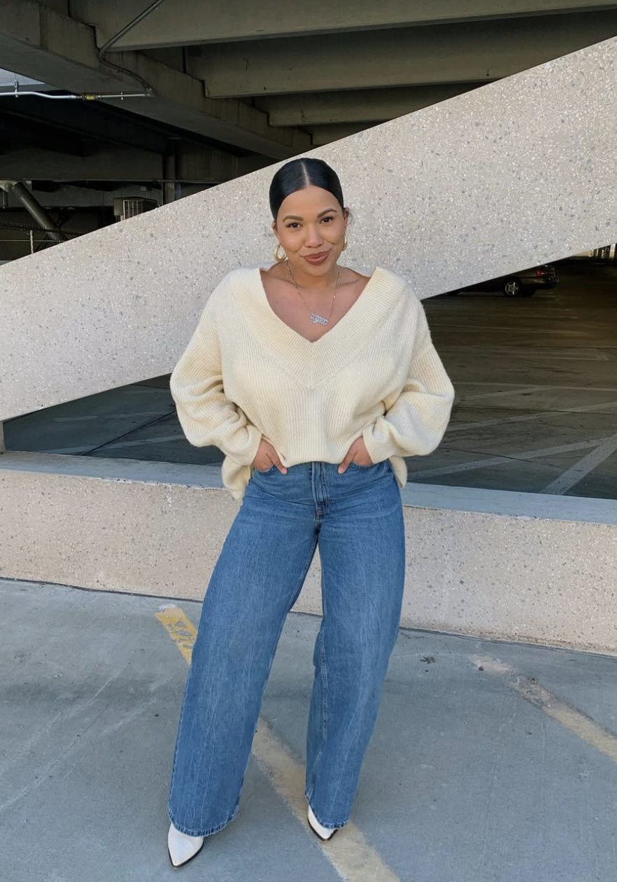 Digital creator Ty Lauren wearing an oversized cream V-neck sweater tucked into medium wash wide-leg jeans, standing in front of an urban concrete backdrop.
