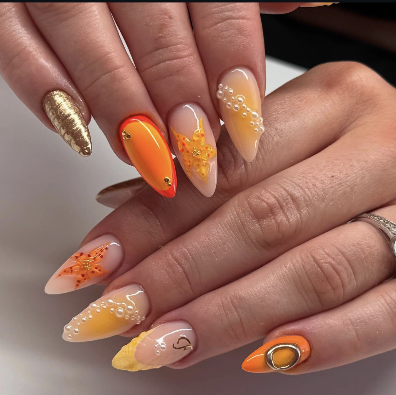 Long almond-shaped nails with a vibrant hot coral glossy finish, featuring decorative orange floral accents and gold leaf details.