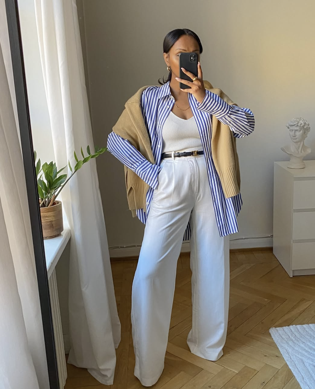 A pro stylist-inspired layering formula featuring a structured white button-down shirt worn under a blue and white striped sweater with light-colored wide-leg trousers.