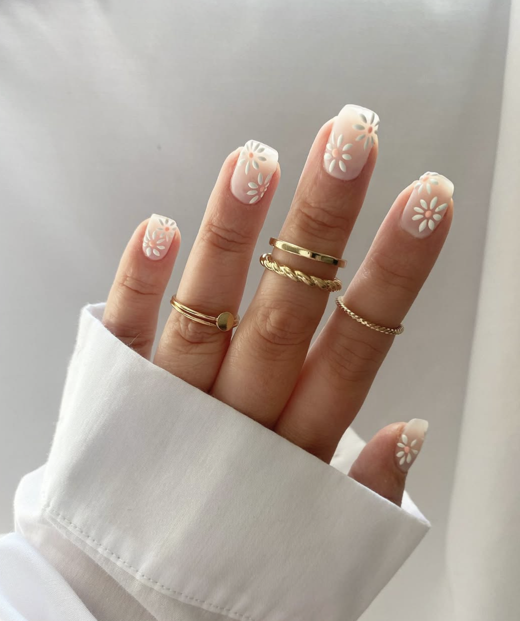 A close-up of square-shaped nails with a sheer nude base, featuring a classic white French manicure decorated with delicate hand-painted tiny orange and white daisy floral patterns, accessorized with stacked gold rings.