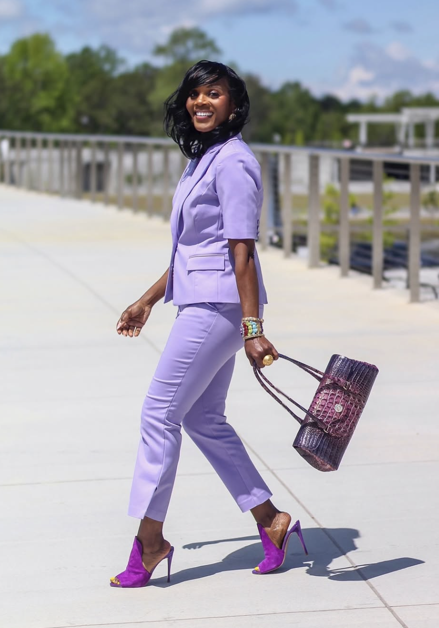 A woman wearing a monochromatic lavender outfit featuring a short-sleeved blazer and matching slim-fit trousers, styled with purple pointed-toe heels and a textured handbag.