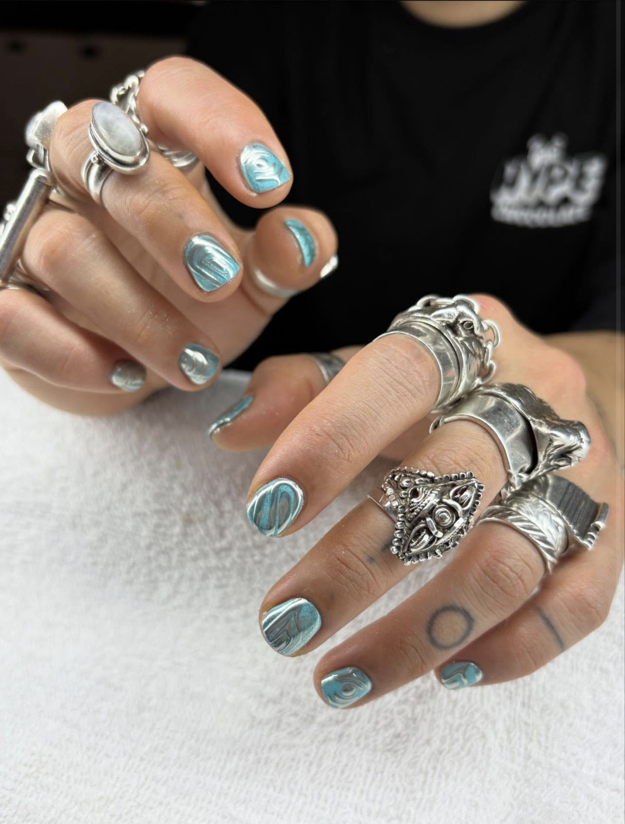 A close-up of square-shaped nails featuring a bright turquoise and metallic silver-blue chrome abstract wave design, heavily accessorized with chunky silver statement rings.