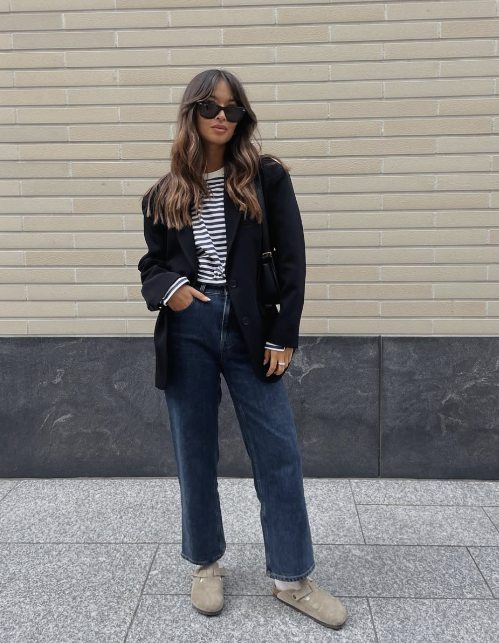 A woman wearing an oversized black blazer over a striped tee and dark-wash straight-leg jeans, accessorized with black sunglasses.